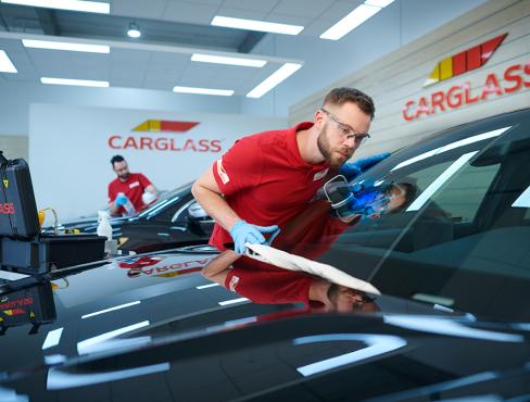  Twee technici in rode shirts en blauwe handschoenen werken aan een autoruit in een Carglass-werkplaats, wat wijst op reparatie van autoruitschade.