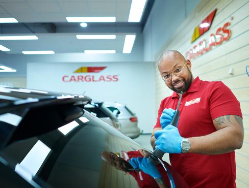 Autotechnicus in rood shirt vervangt ruitenwisser in werkplaats, met CARGLASS-logo op achtergrond.