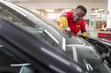 Autotechnicus in rood uniform en gele handschoenen repareert een autoruit in een Carglass-werkplaats, met rode gereedschapskasten op de achtergrond.